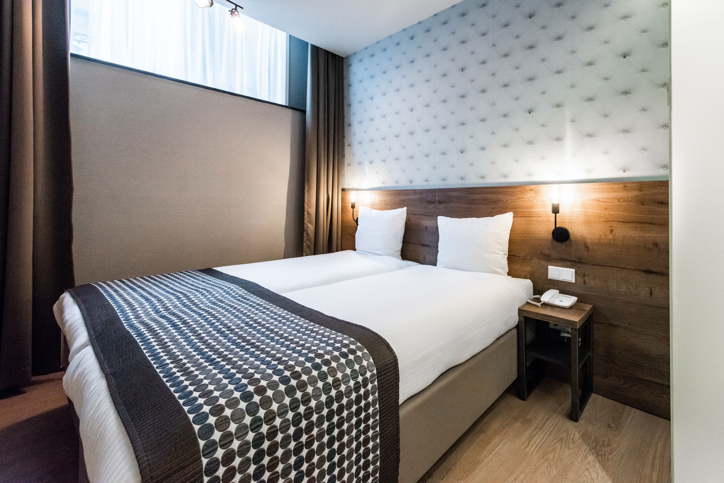 A hotel room with a large bed, a wooden headboard, a bedside table with a phone, and a window with curtains.