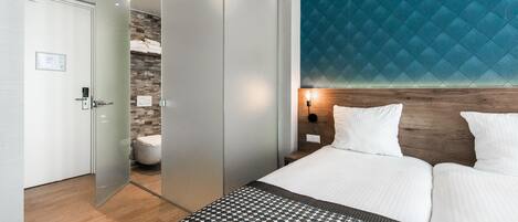 Una habitación de hotel moderna con una cama grande, una puerta de vidrio que conduce a un baño y una pared azul con textura.