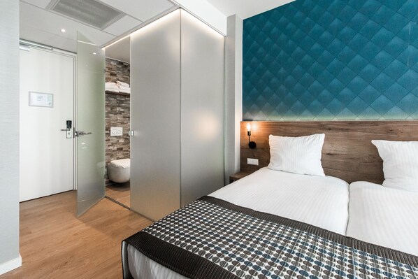 Una habitación de hotel moderna con una cama grande, una puerta de vidrio que conduce a un baño y una pared azul con textura.