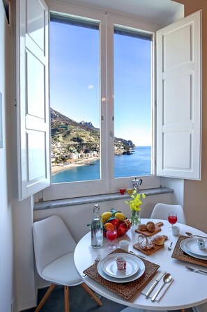 Un comedor con mesa dispuesta para dos, vista al mar y ventana con contraventanas blancas.