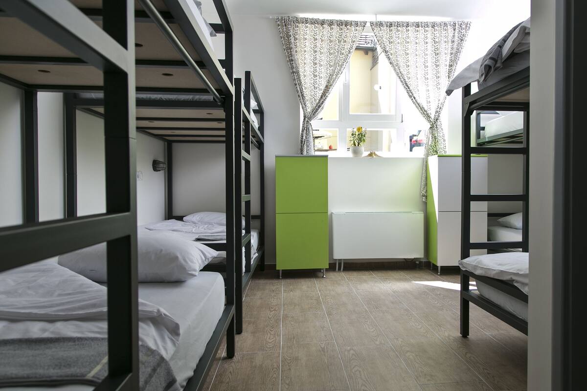 Quarto em dormitório com beliches, uma kitchenette e janela com cortinas.