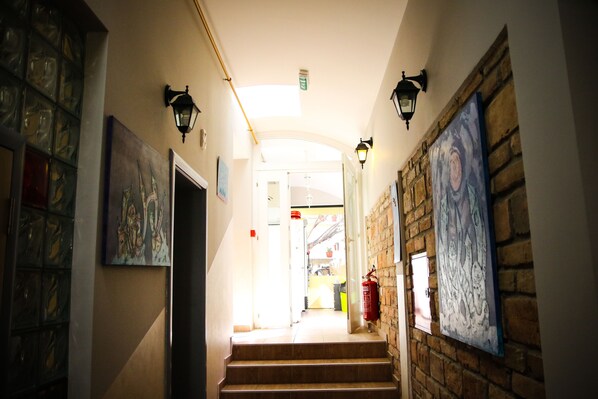 A hallway with a staircase leading to a bright room, featuring wall-mounted artwork and a fire extinguisher. - Varad Inn Hostel and Cafe (Novi Sad)