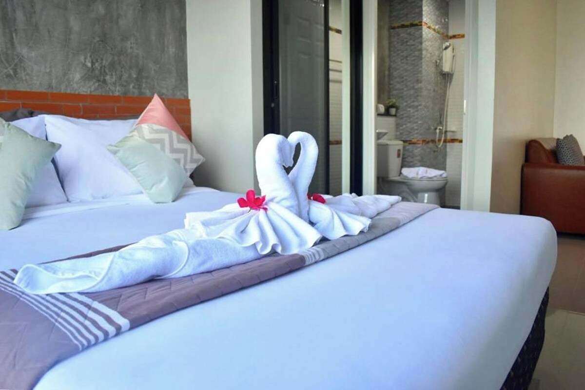 Un lit bien fait, agrémenté d’une décoration en forme de cygnes réalisée avec des serviettes, une tête de lit en briques et une salle de bain où l’on peut voir une douche et des toilettes.