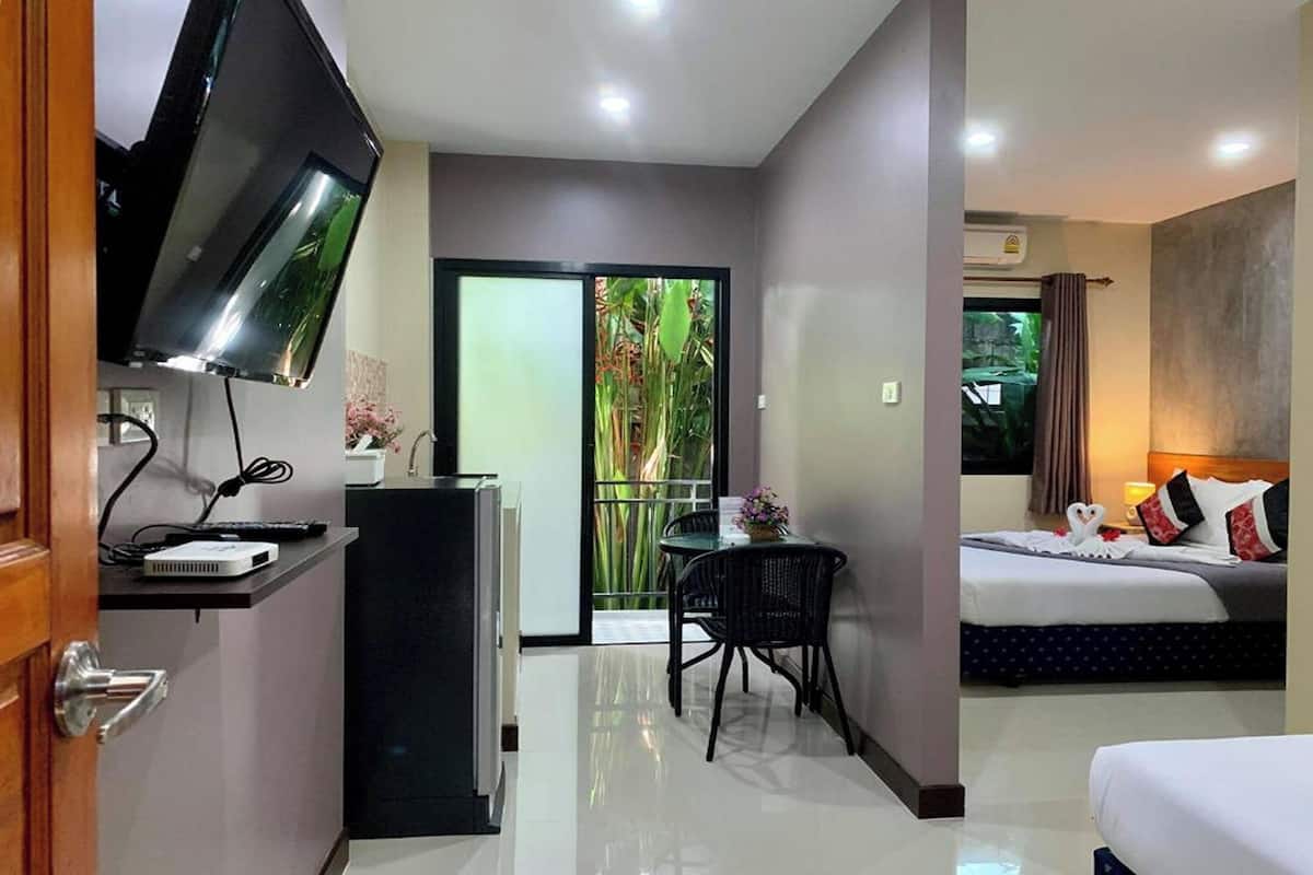A hotel room with a bed, a desk with a TV, a small table with chairs, and a balcony with plants.