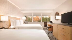 Habitación de hotel con dos camas, un escritorio, un televisor y una ventana grande con vistas a follaje otoñal.