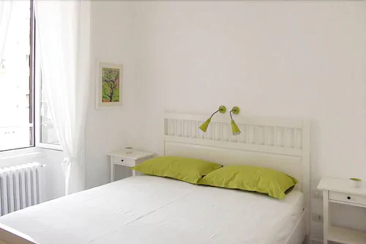 A white bed with green pillows, a white nightstand, a white bedside table, and a window with white curtains.