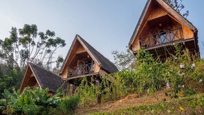 Three wooden cabins with triangular roofs, surrounded by lush greenery and banana plants. - Rwiza Village (Mubuga)