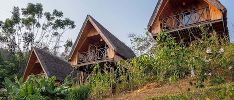 Trois cabanes en bois, dotées de toits triangulaires, sont entourées d’une végétation luxuriante et de bananiers.