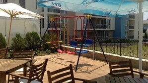 An outdoor patio area with a swing set, a table with chairs, and a large umbrella. - Tropics Hotel (Sveti Vlas)