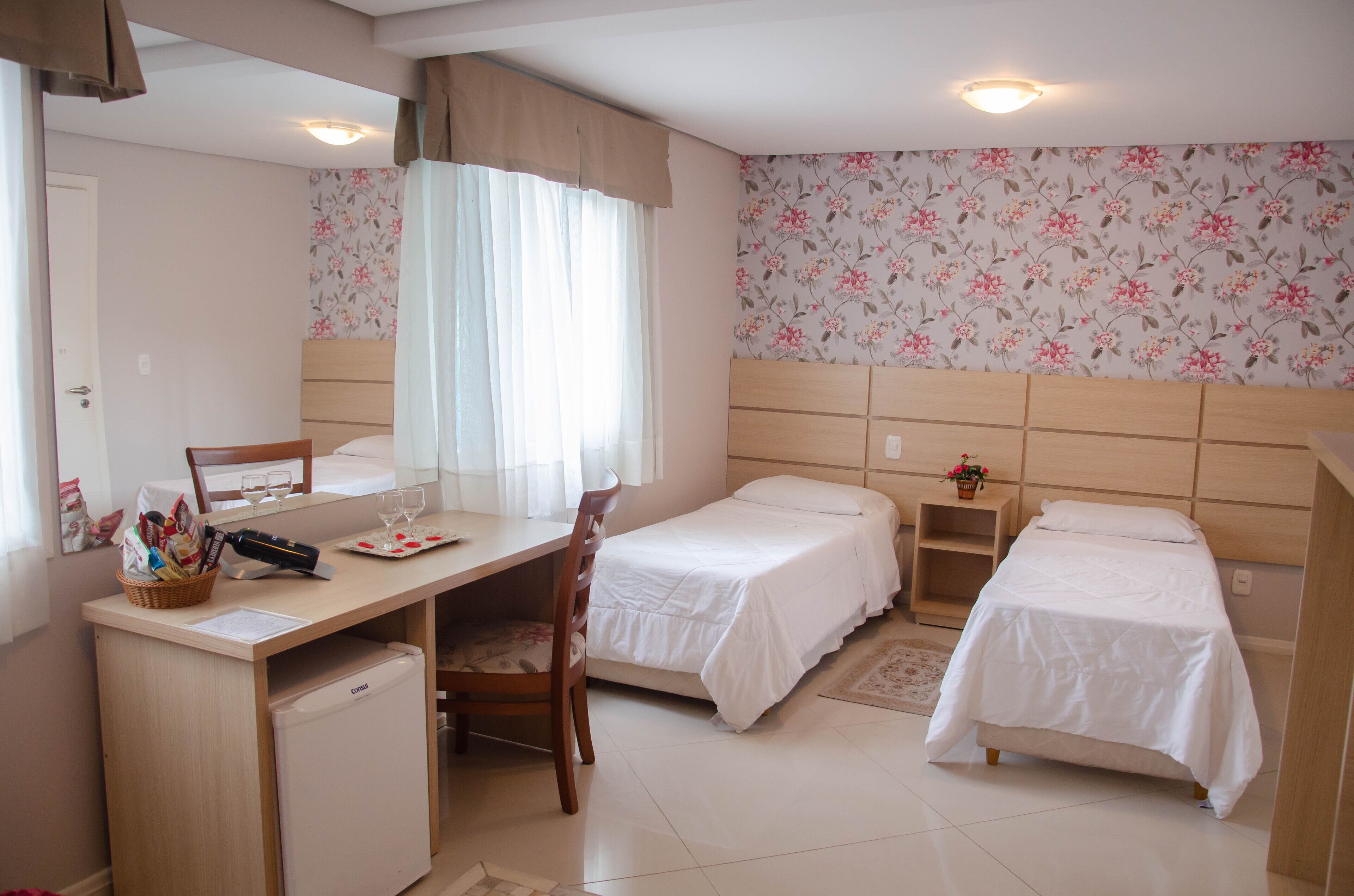 A hotel room with a desk, two single beds, a small refrigerator, and a window with curtains.