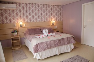 Una cama bien tendida con colcha rosa y blanca, dos lámparas de noche, una mesita de noche con un jarrón de flores y un aire acondicionado instalado en la pared.