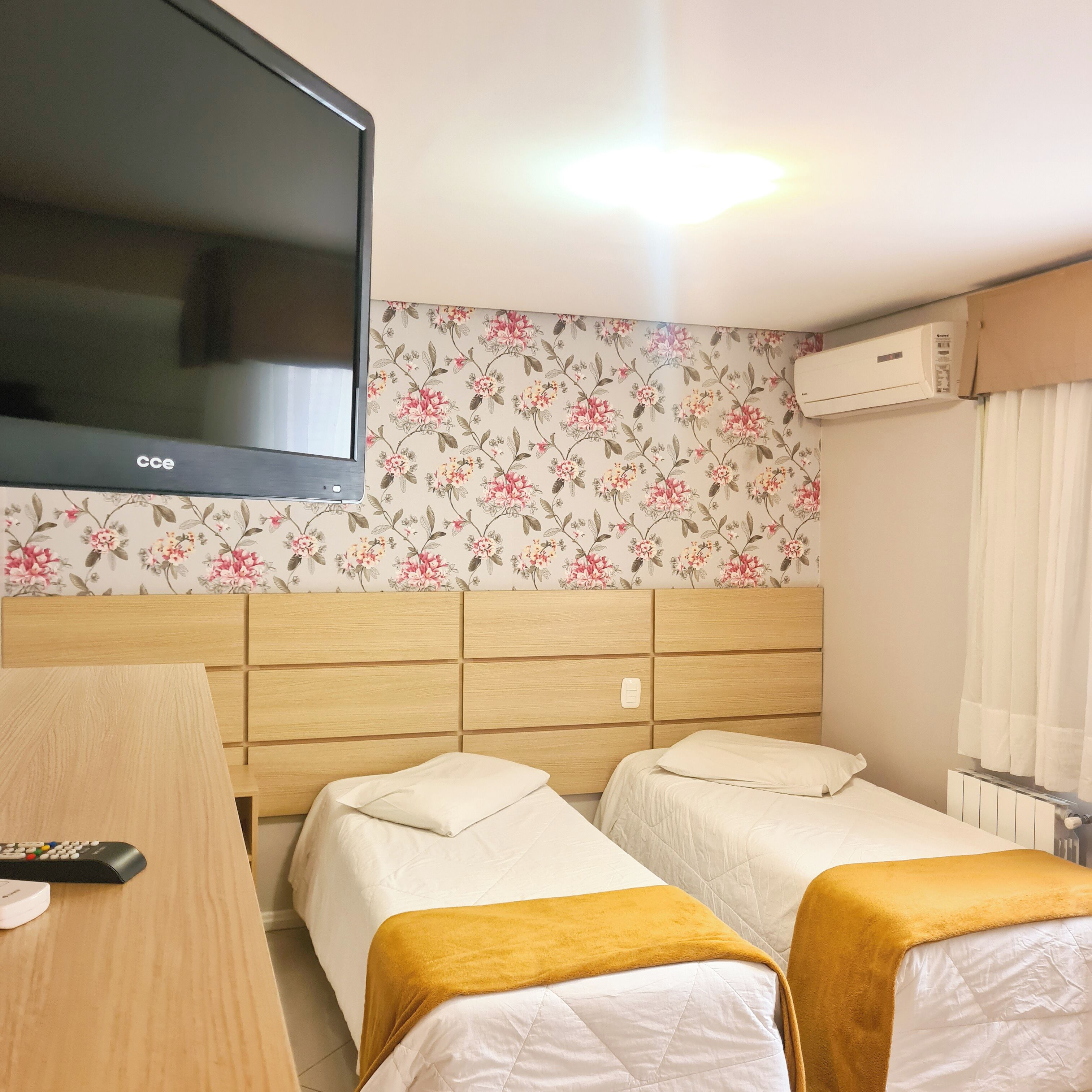 A hotel room with two single beds, a wooden headboard, a flat-screen TV, floral wallpaper, an air conditioning unit, and a white curtain.