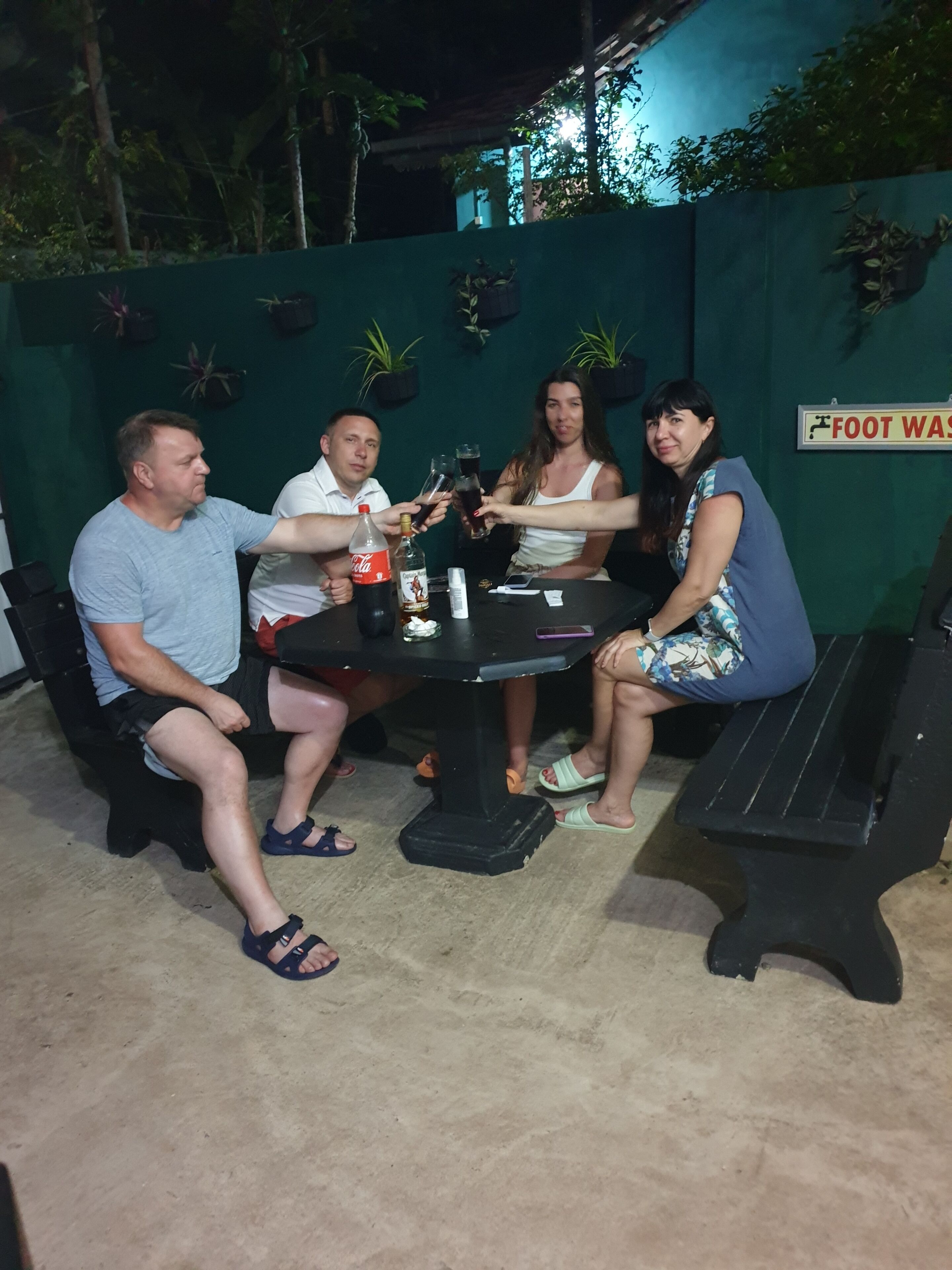 Un grupo de personas sentadas alrededor de una mesa con bebidas, en un espacio al aire libre con un muro verde y plantas.