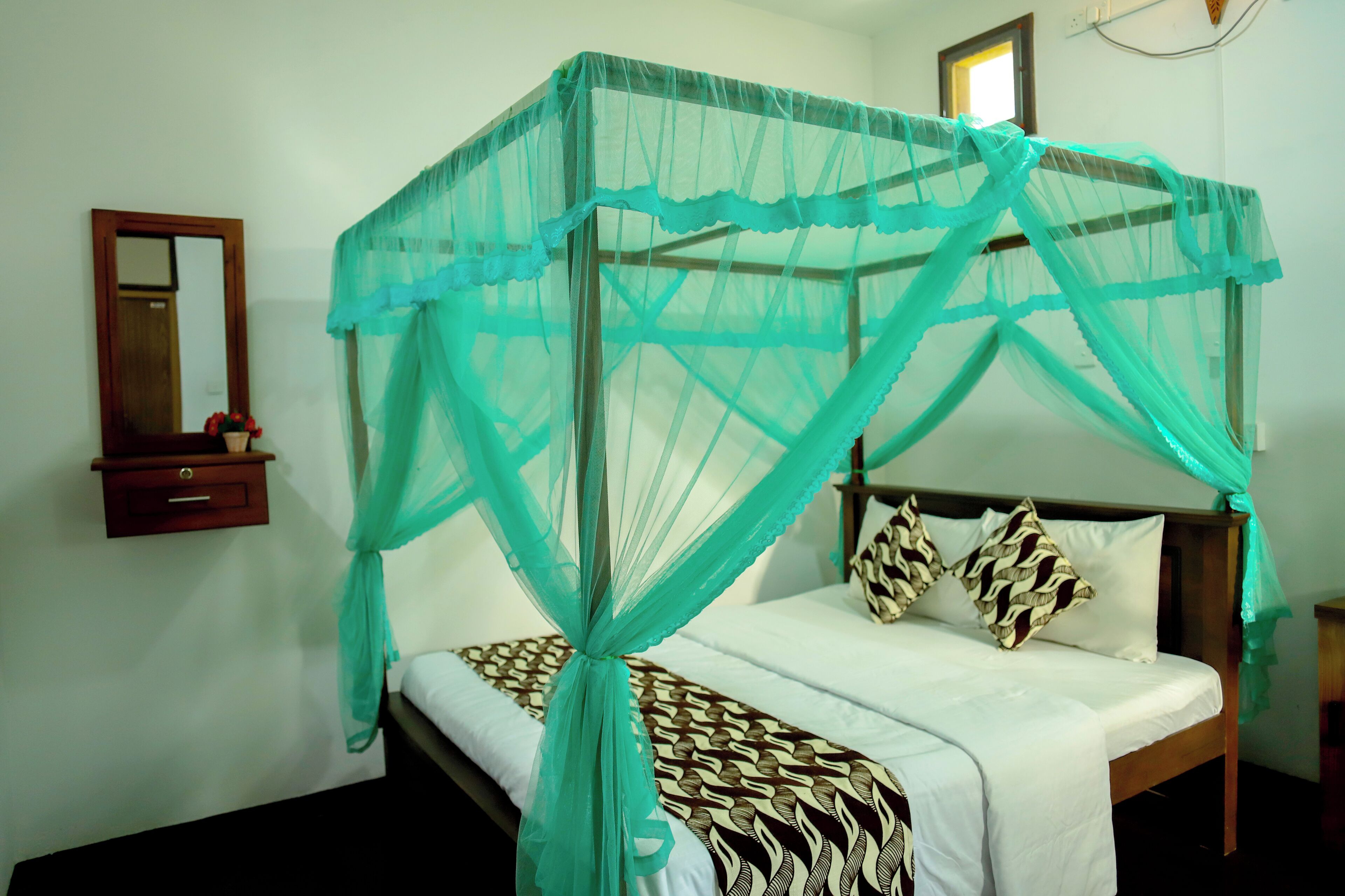 A bed with a mosquito net, a bedside table, a mirror, and a wall-mounted shelf with a vase.