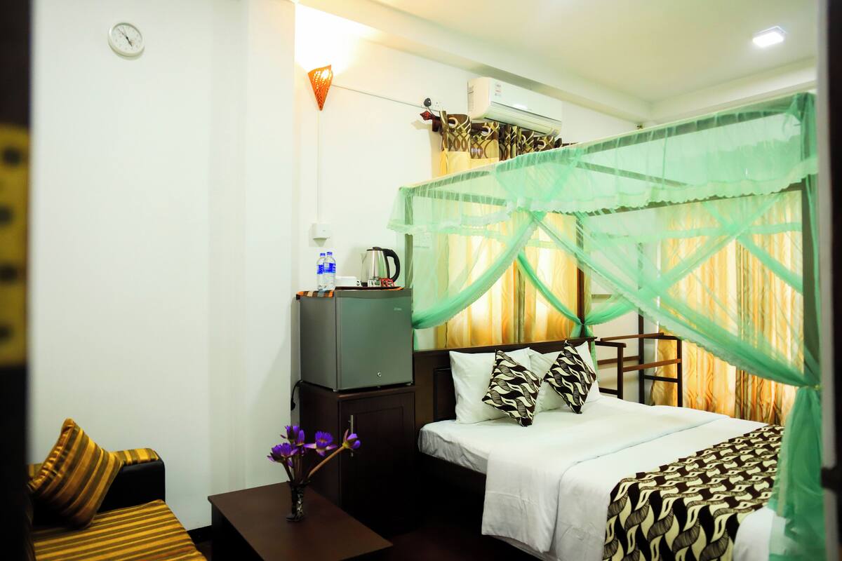 A hotel room with a bed, a nightstand, a small table with a vase of flowers, a heater, and a window with a mosquito net.