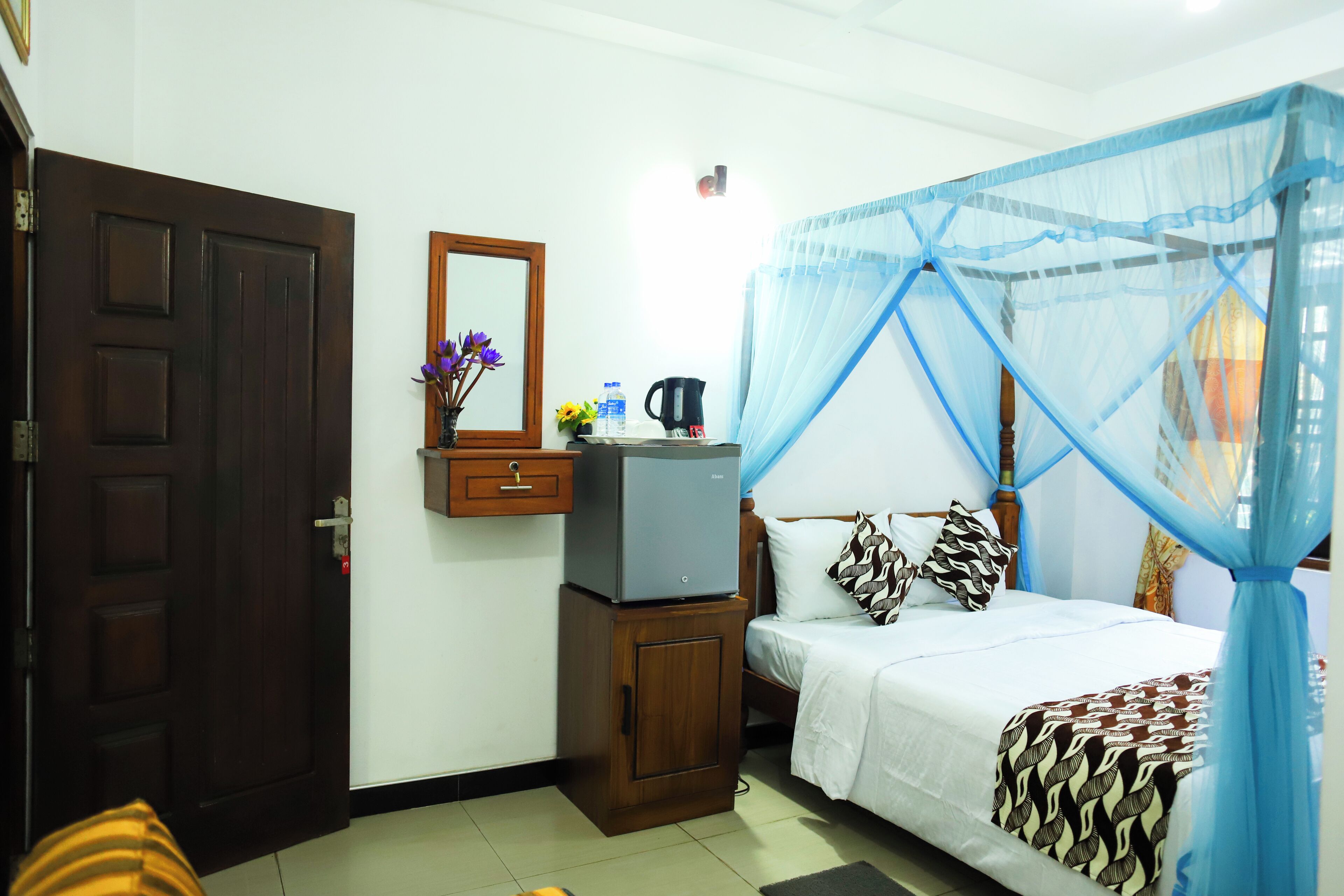 A bedroom with a bed, a nightstand, a mirror, a wooden cabinet, and a blue mosquito net.