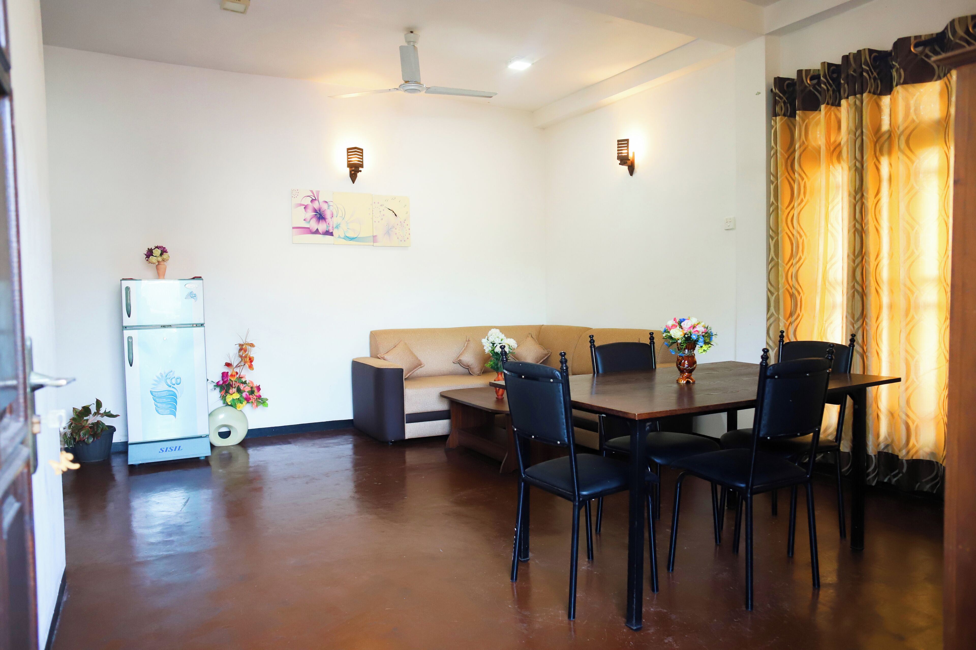 A dining area with a table, chairs, a refrigerator, a sofa, and wall-mounted lights.