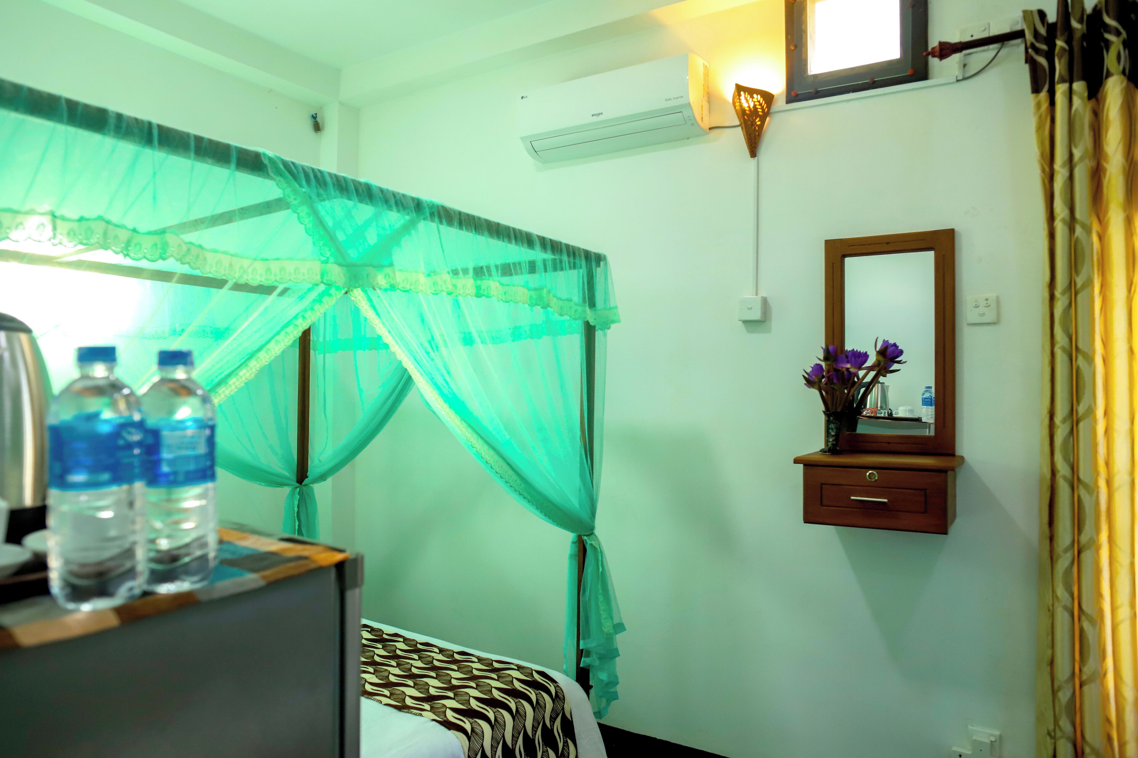 Un dormitorio con una cama, una mesita de noche con botellas de agua, una mesilla, un espejo y un aire acondicionado instalado en la pared.