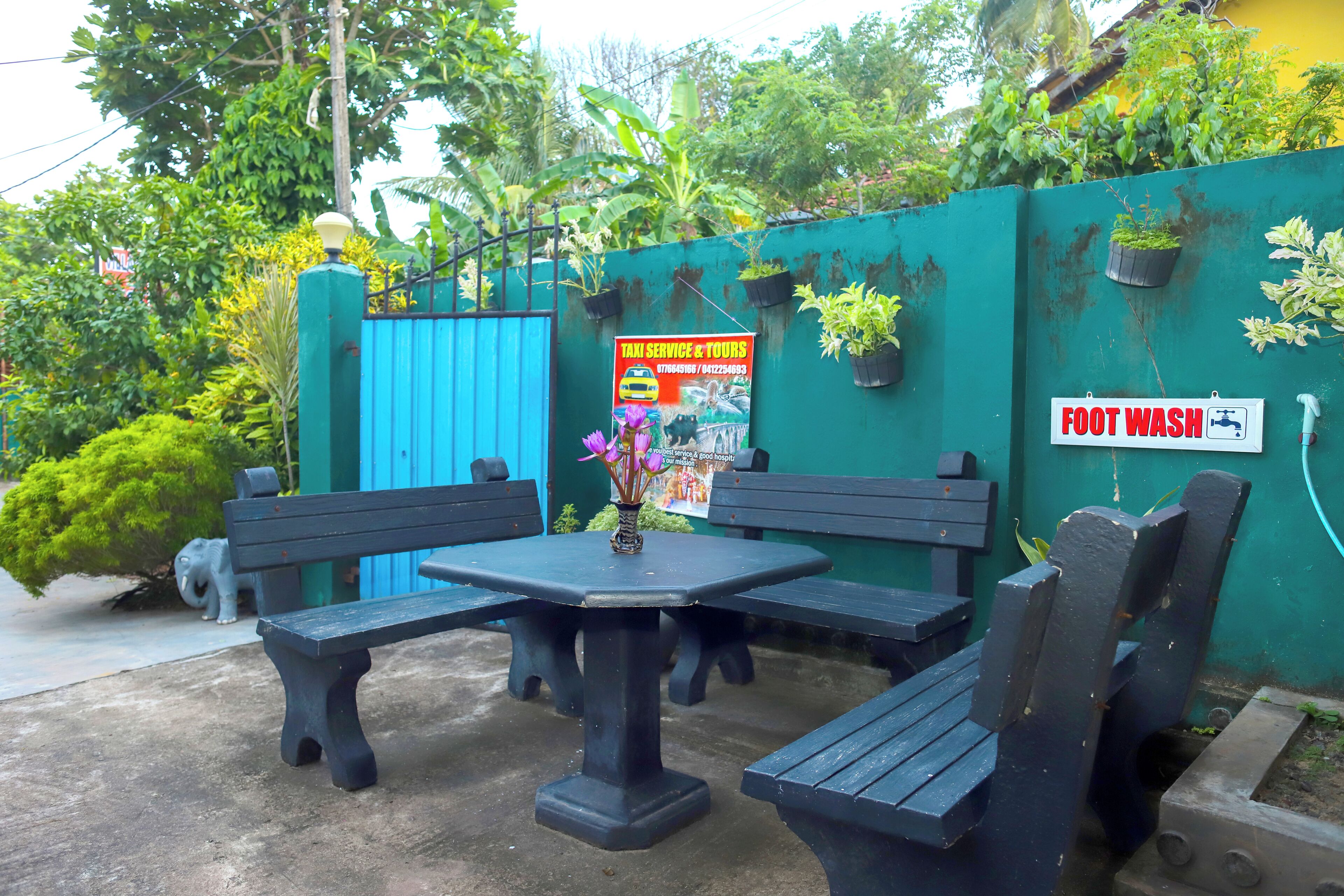 An outdoor seating area with black benches and a table, surrounded by a green wall with potted plants and a blue gate.