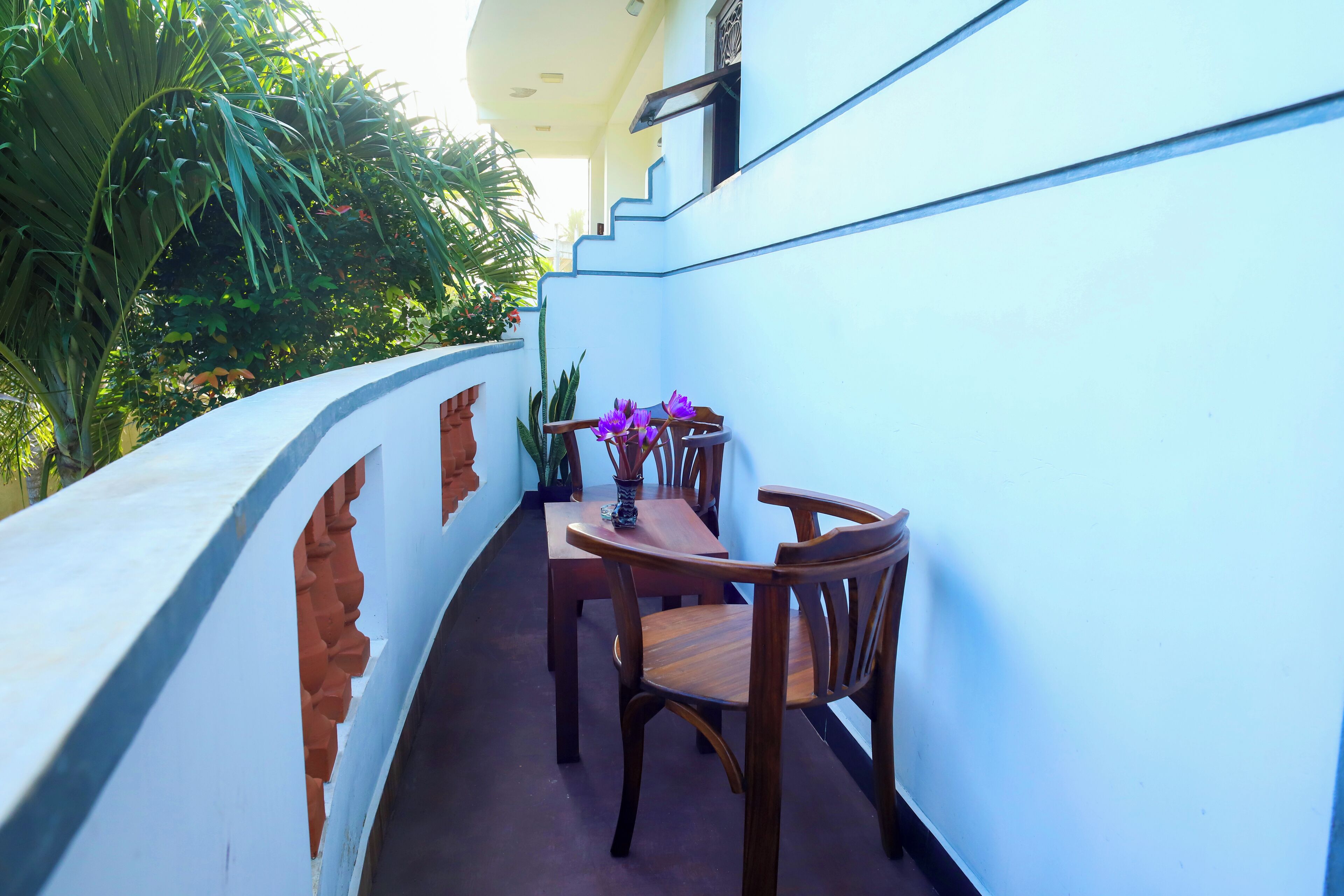 A narrow balcony with a wooden table and chairs, a vase with purple flowers, and a view of lush greenery.