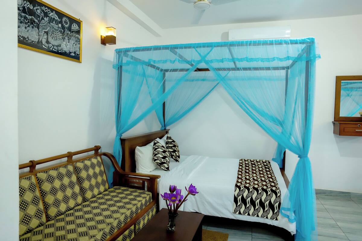 A room with a bed under a blue mosquito net, a sofa with patterned cushions, and a wall-mounted light fixture.