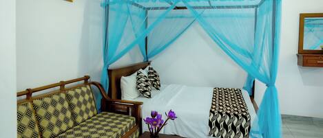A room with a bed under a blue mosquito net, a sofa with patterned cushions, and a wall-mounted light fixture.