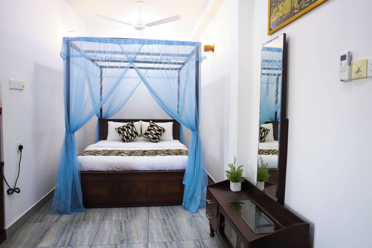 A bedroom with a bed under a blue canopy, a wooden headboard, a small bedside table with a plant, and a mirror on the wall.