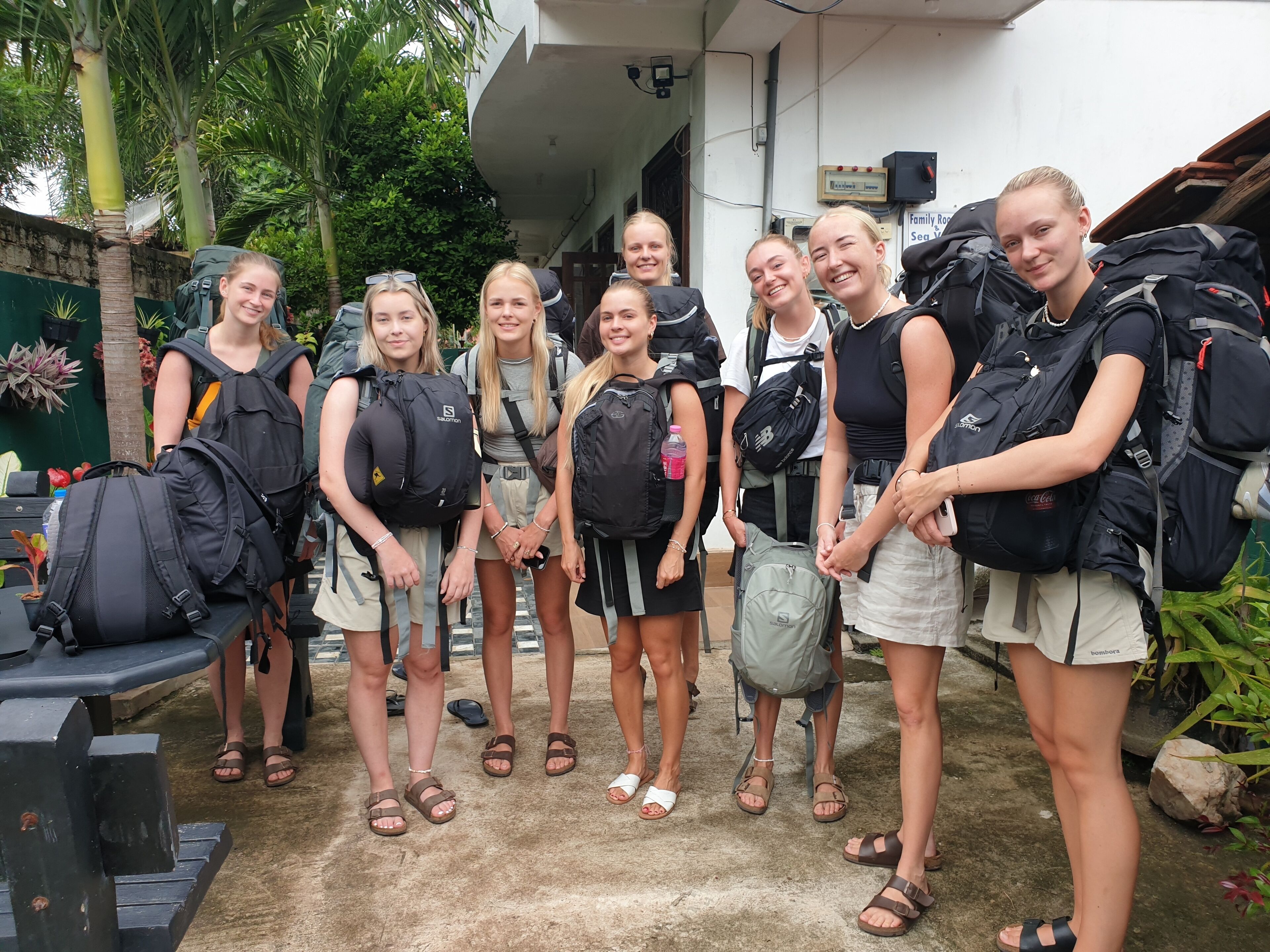 Un groupe de personnes, portant des sacs à dos, se tient dans un espace extérieur doté d’un sol en béton et dont l’arrière-plan est composé de verdure.