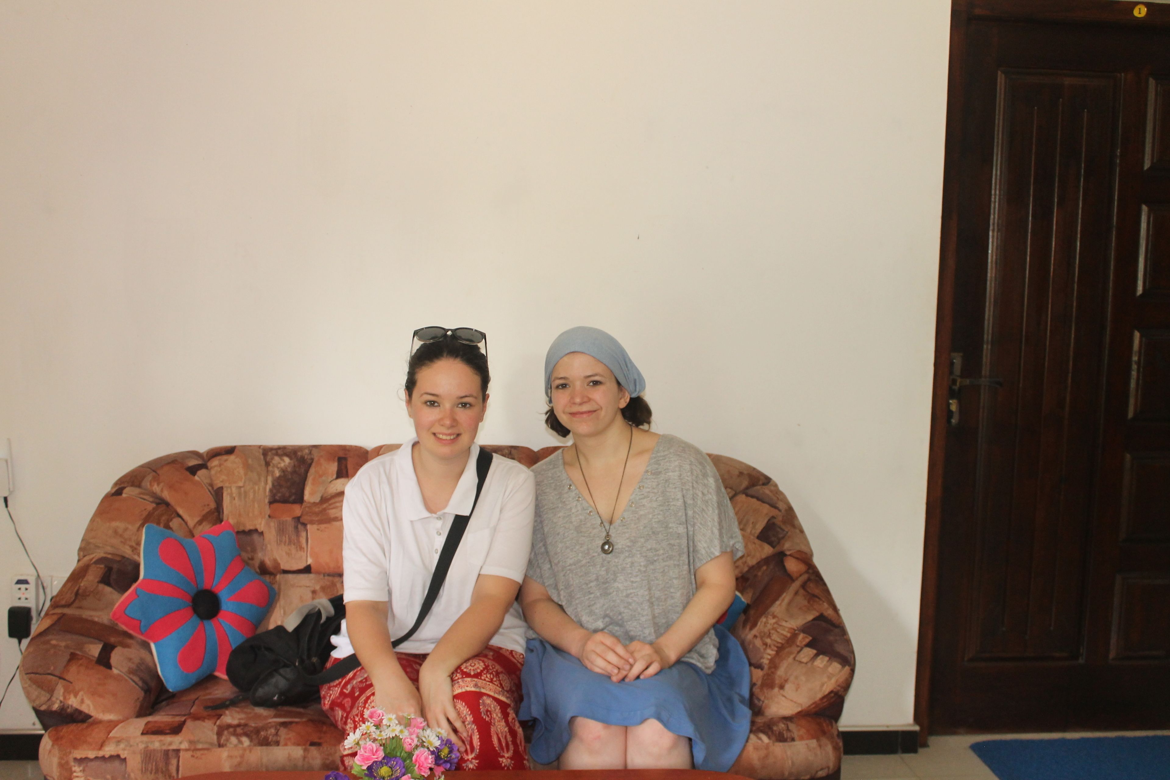 Two people sitting on a brown couch with a colorful pillow and a black bag.