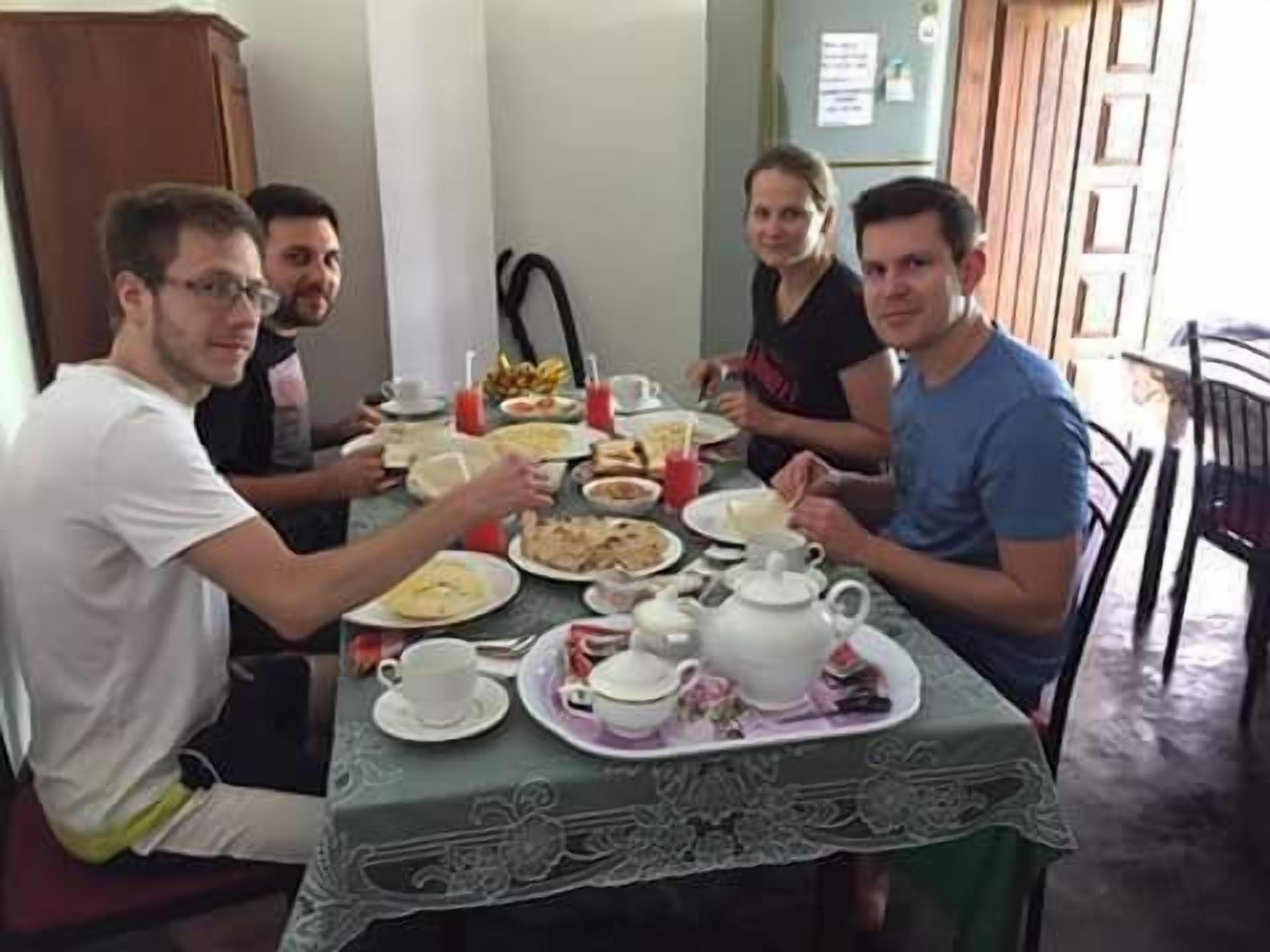 A group of people seated around a table with plates of food, cups, and a teapot, indicating a meal setting.
