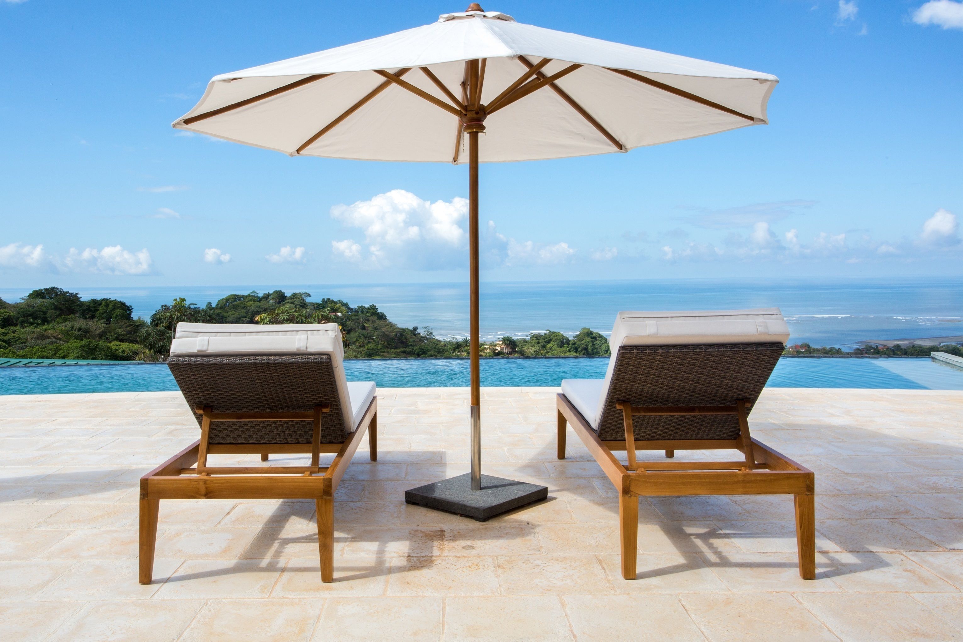 Two lounge chairs with a large umbrella, a pool, and a view of the ocean and greenery.