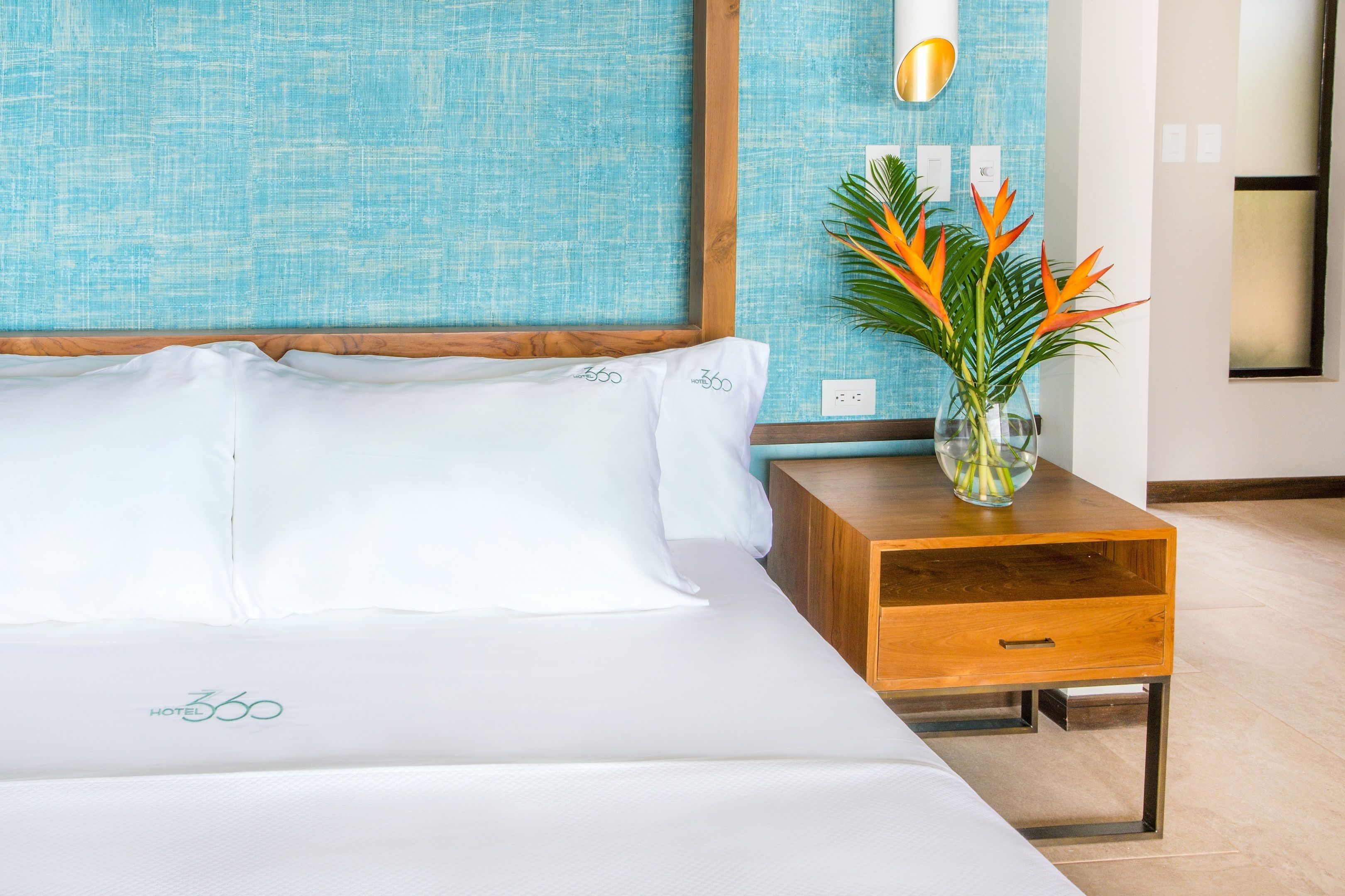 A hotel room with a bed, a wooden nightstand, a vase with flowers, and a blue textured wall.