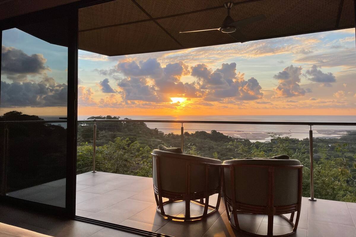 A balcony with two wicker chairs facing a sunset view over a coastal landscape.