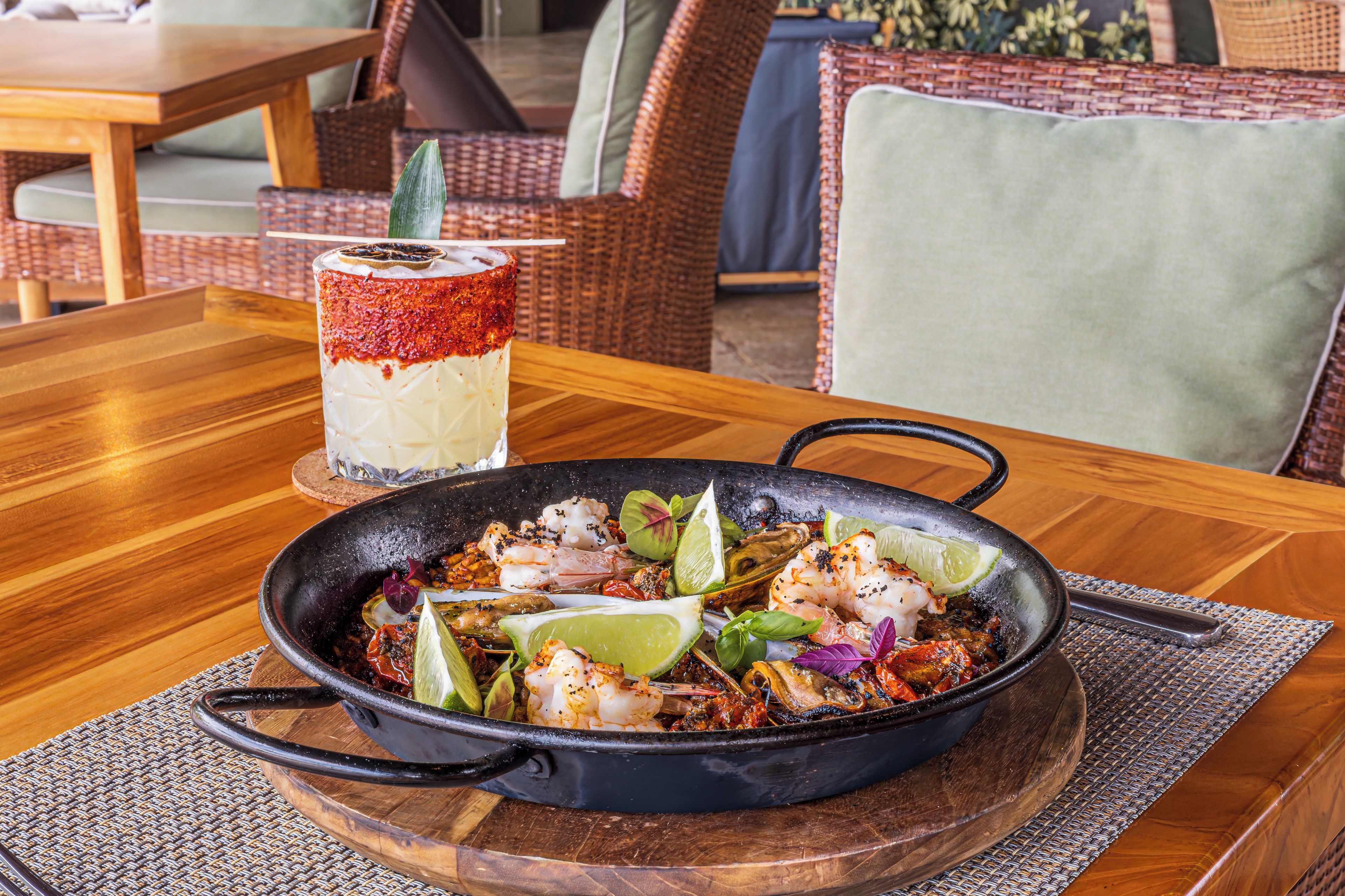A dining table with a skillet of seafood, a glass of a layered drink, and wicker furniture in the background.