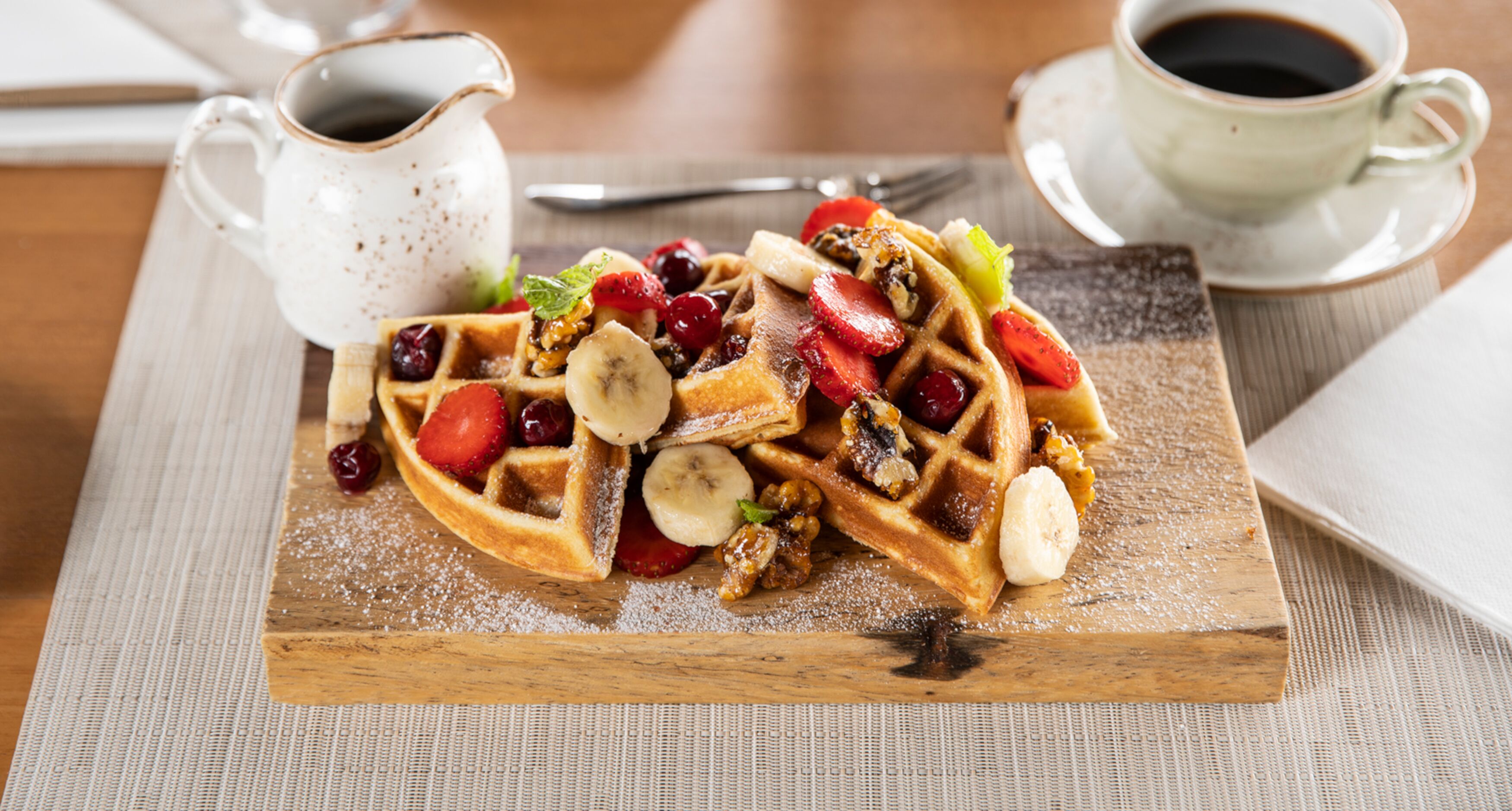 A breakfast plate with waffles topped with strawberries, bananas, and nuts, accompanied by a cup of coffee and a jug of syrup.