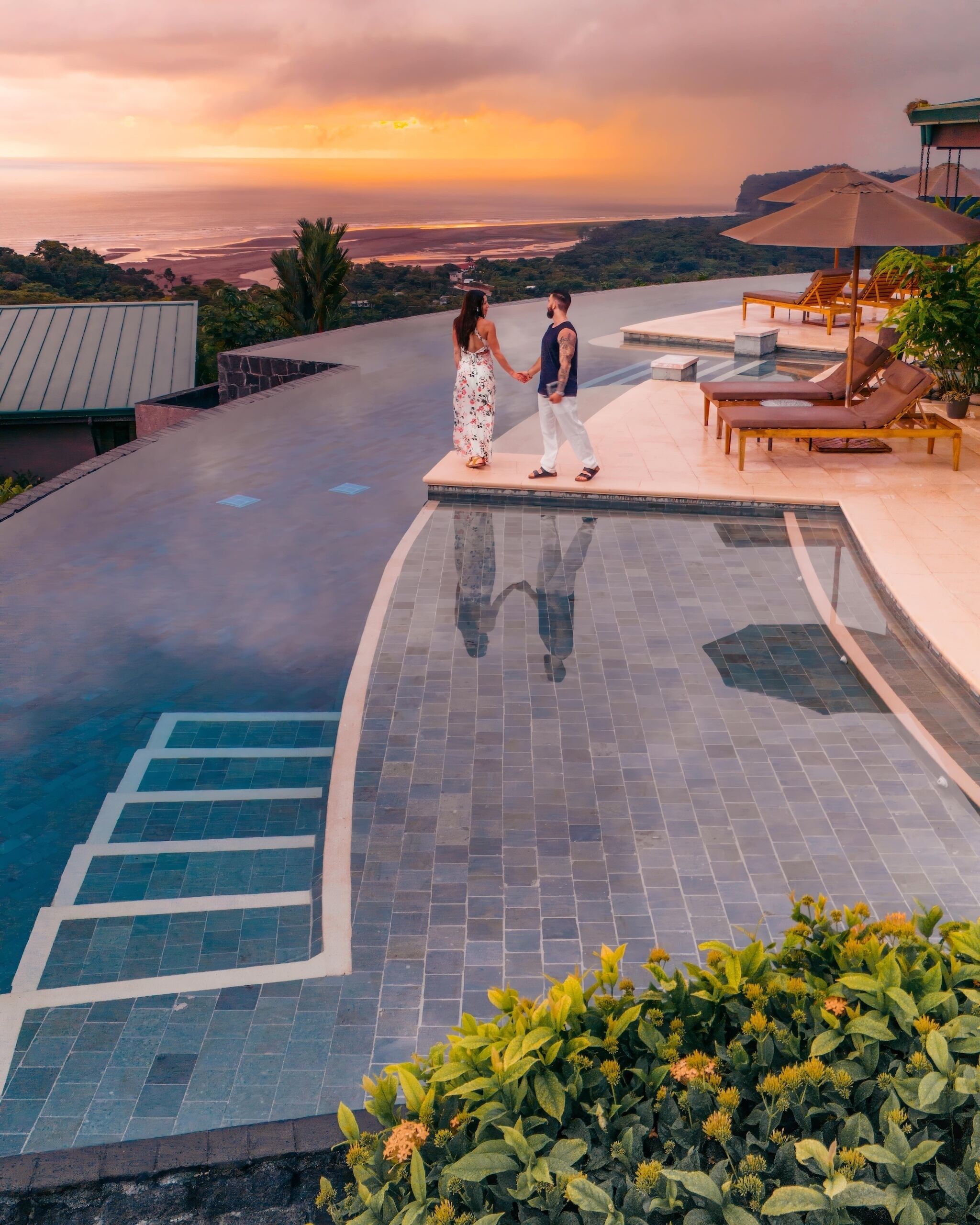 Ein Paar, das Händchen haltend auf einer Poolterrasse mit Blick auf das Meer und einen Sonnenuntergang steht.