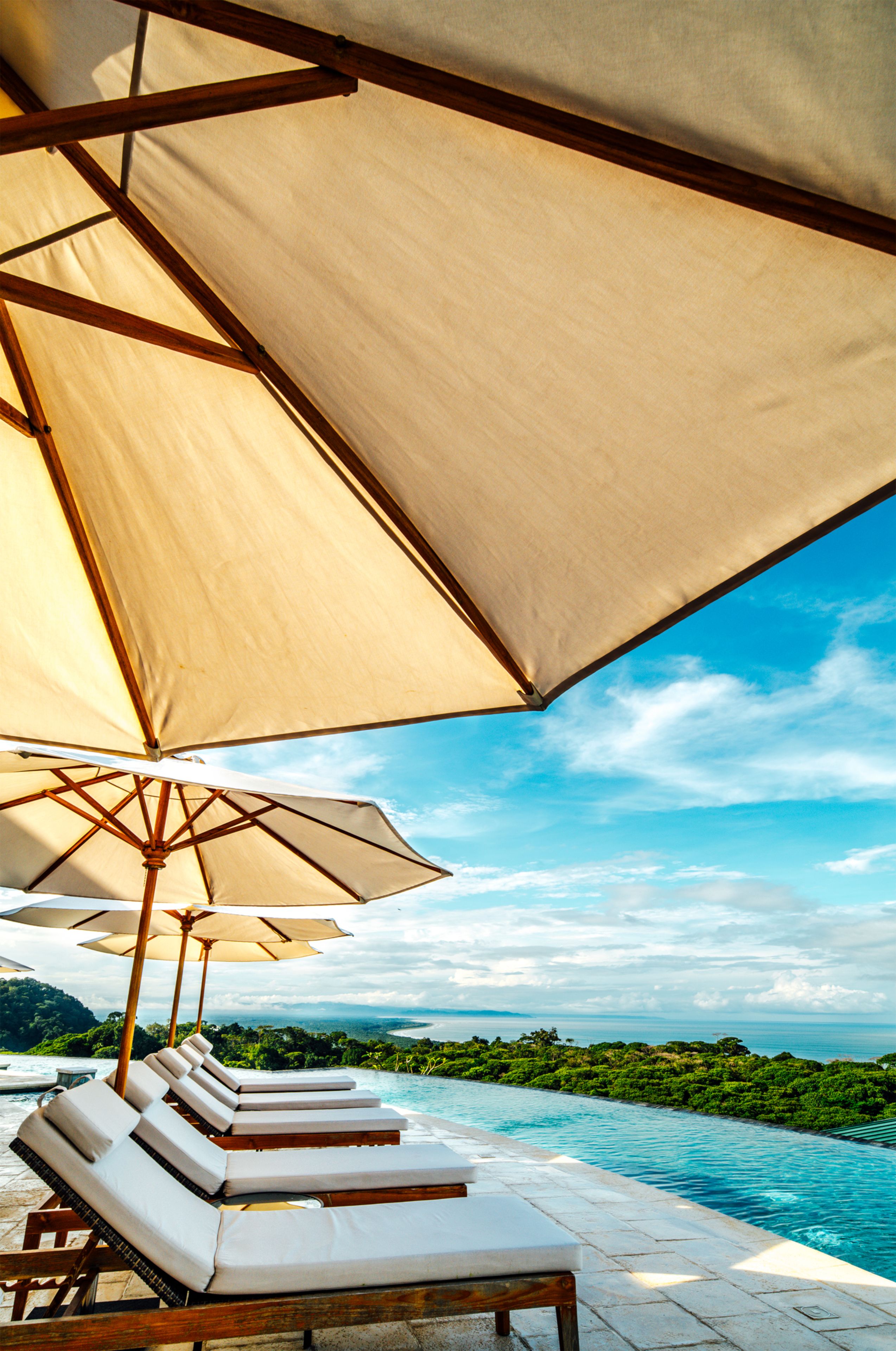 A poolside area with lounge chairs and umbrellas, overlooking a clear blue sky and a lush green landscape.