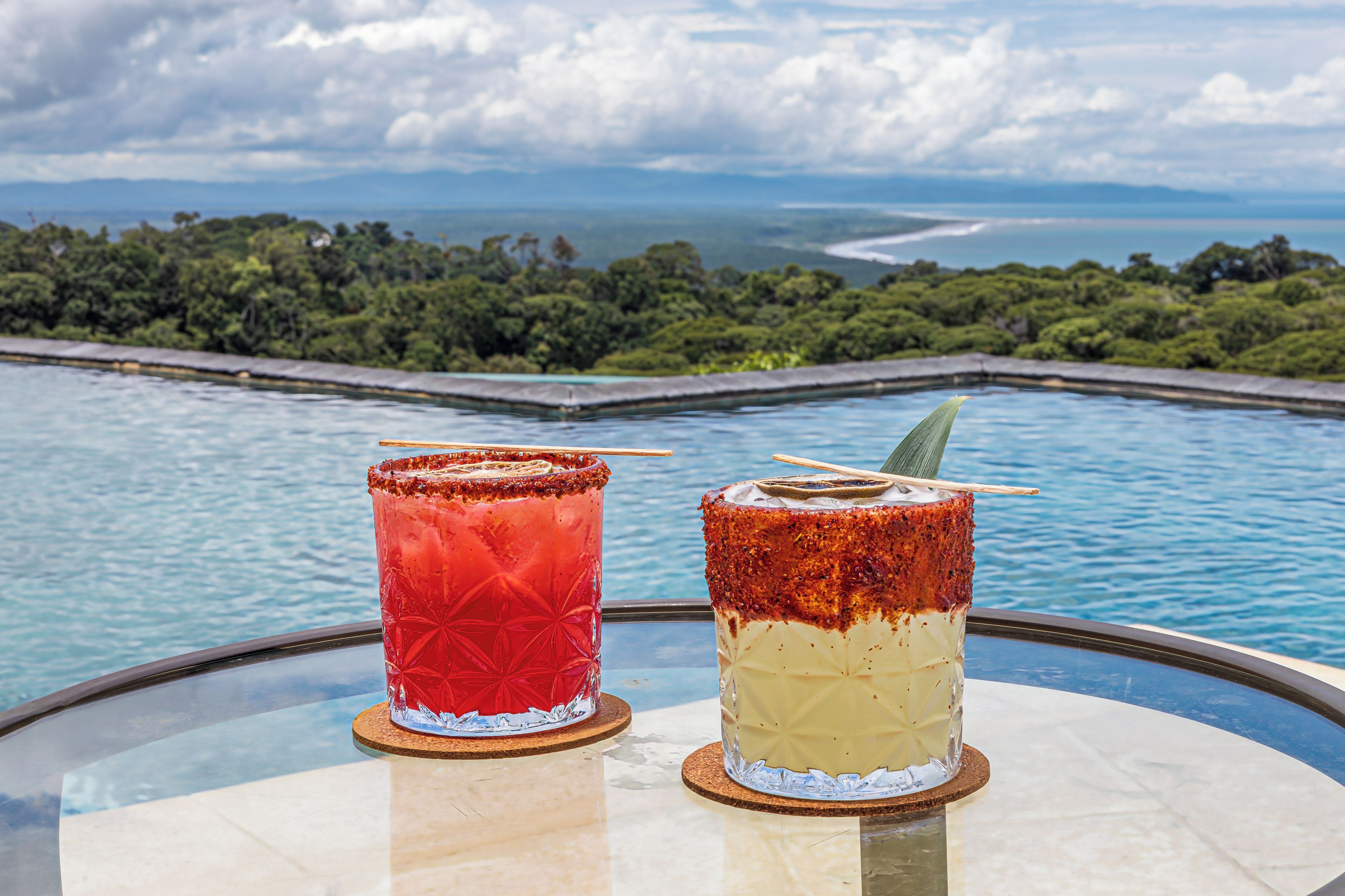 Two drinks on a glass table with a view of a body of water and greenery in the background.