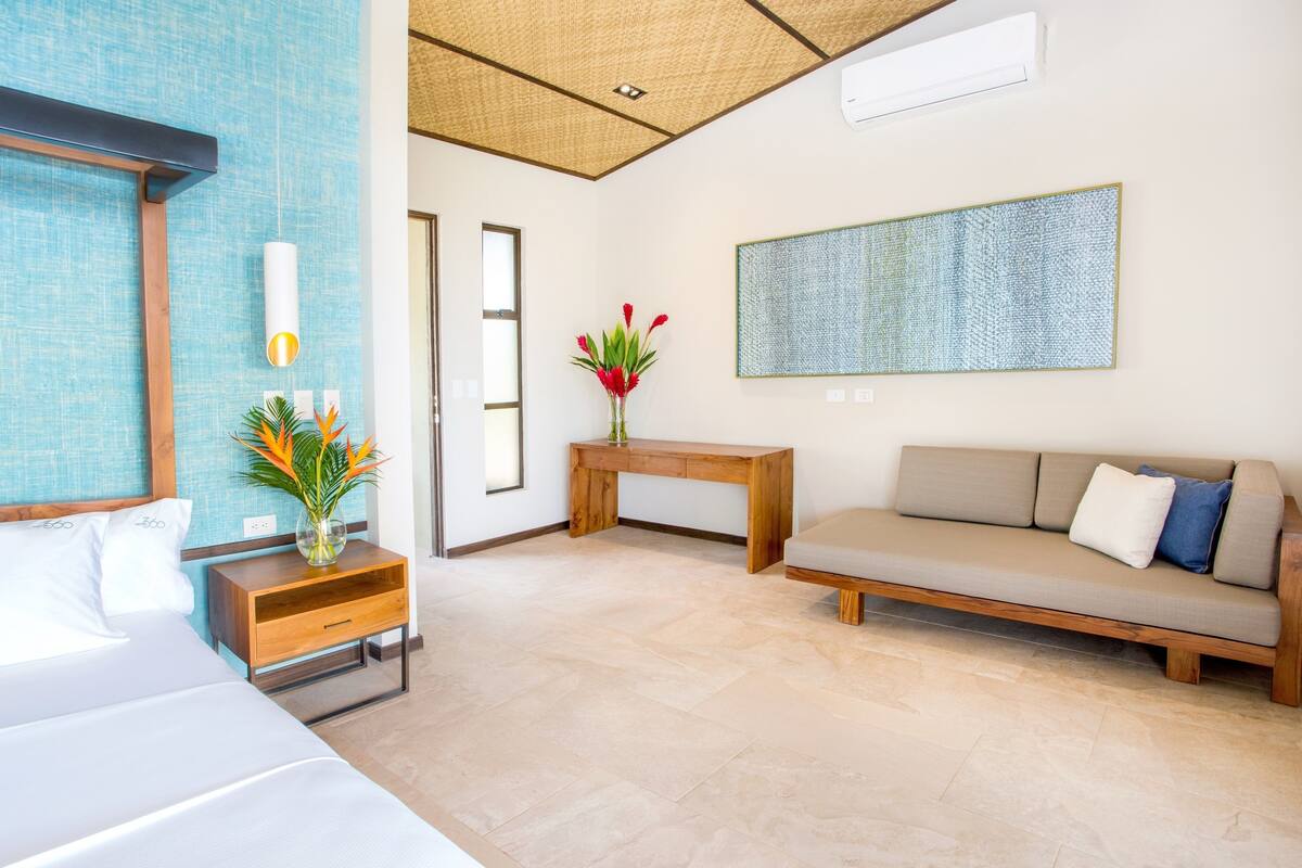A modern hotel room with a bed, a sofa, a wooden nightstand, a vase with flowers, a small table, and a large abstract painting on the wall.