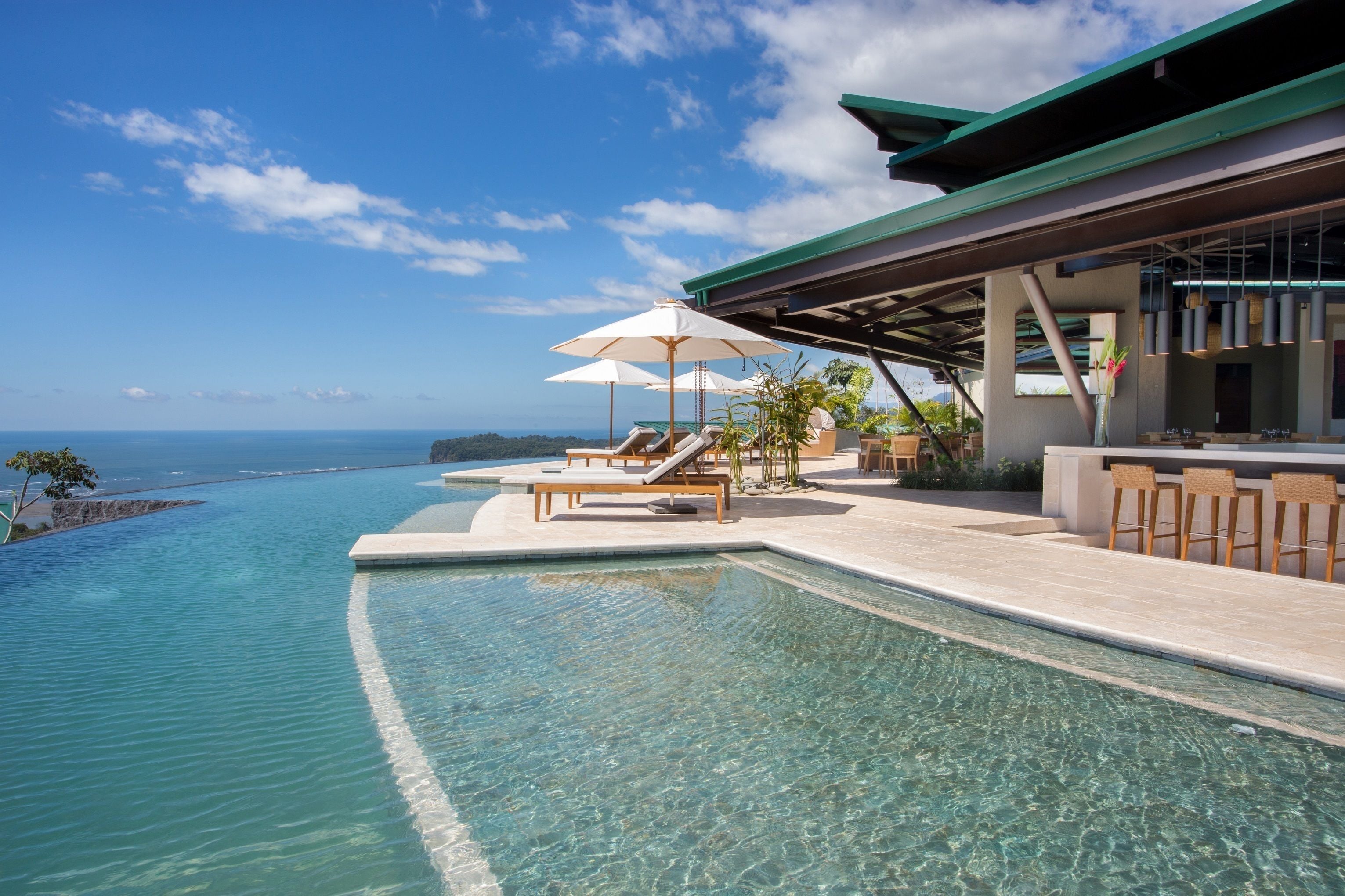 A pool with clear water, lounge chairs, umbrellas, and a bar area with seating.