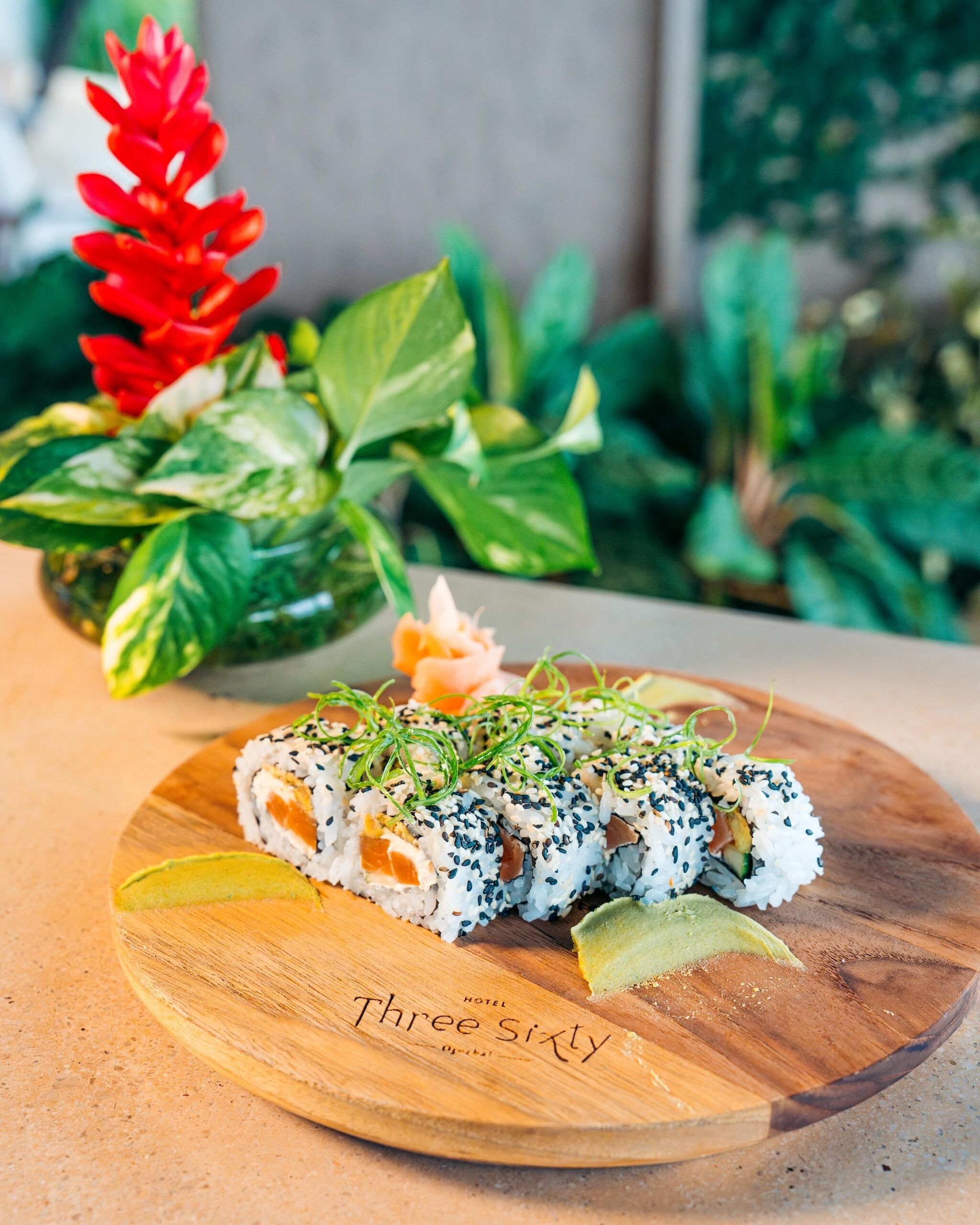 A wooden plate with sushi rolls garnished with green herbs and sesame seeds, accompanied by a slice of avocado and a small orange garnish.