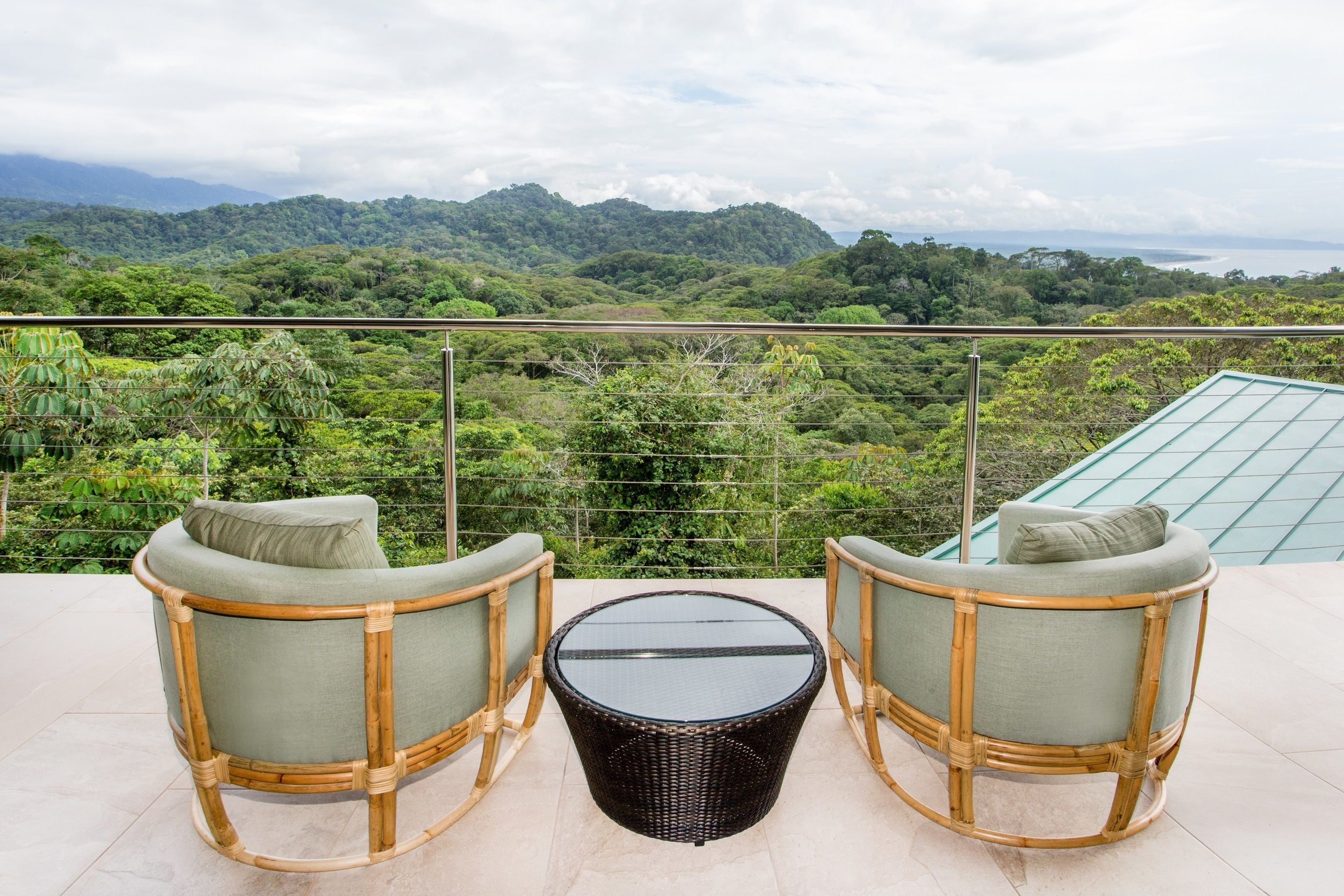 Un balcón con dos sillones de mimbre y una mesa de vidrio, con vistas a un paisaje verde exuberante y montañas.