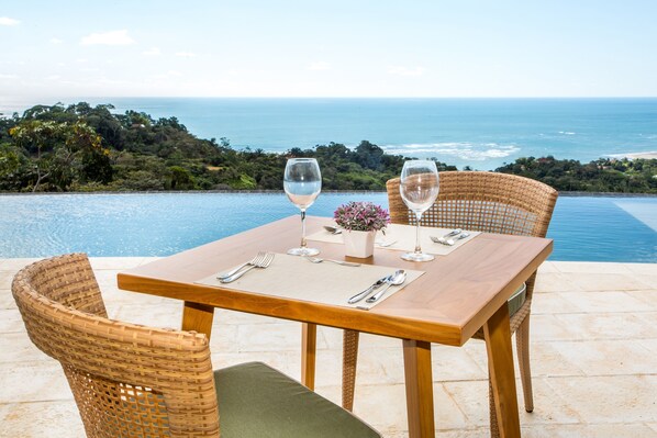 Mesa de comedor con dos sillas, preparada con copas de vino, cubiertos y un florero, con vista a una piscina y al océano.
