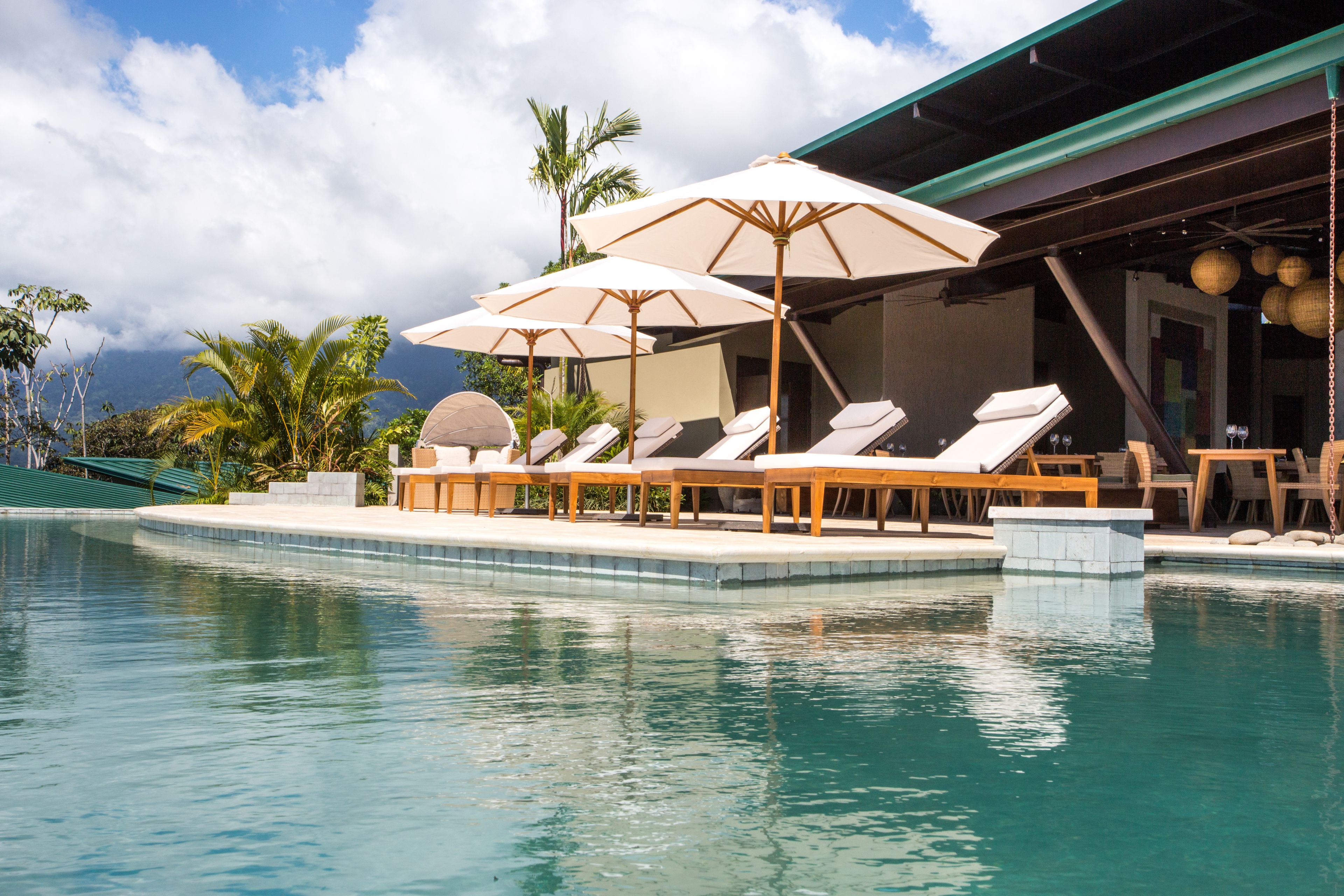 A pool area with lounge chairs and umbrellas, surrounded by palm trees and a building with a green roof.