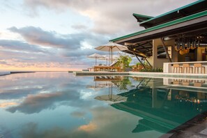 Une piscine à débordement dotée de chaises longues et de parasols, dont la surface reflète le ciel et les nuages.
