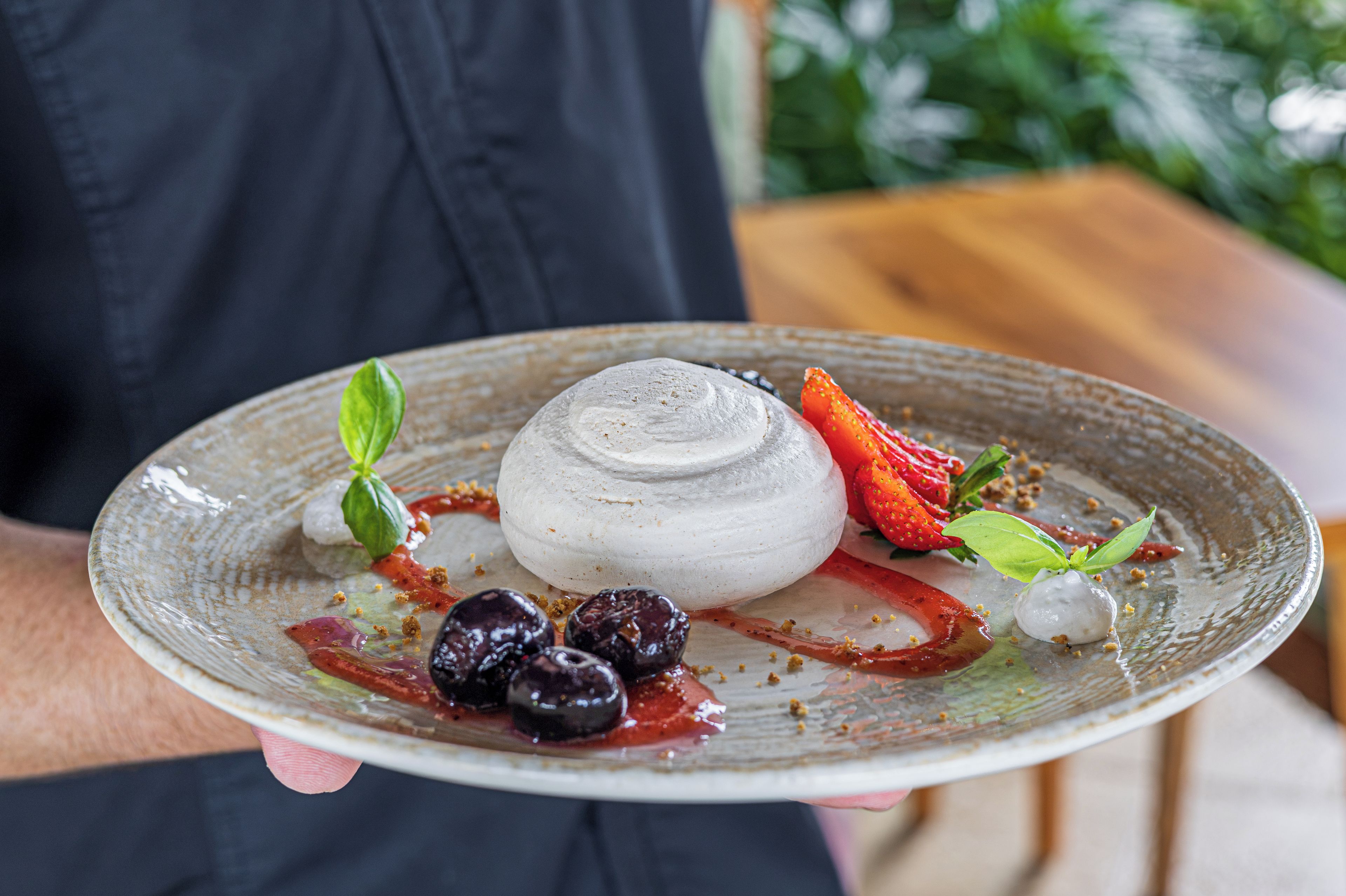 A person holding a plate with a dessert that includes a round white item, strawberries, and a basil leaf.