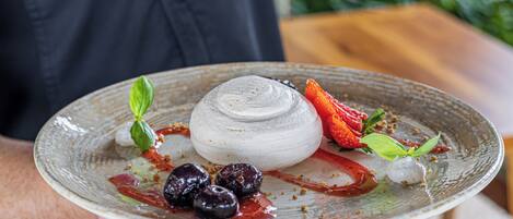Una persona sostiene un plato con un postre que incluye un elemento blanco redondo, fresas y una hoja de albahaca.
