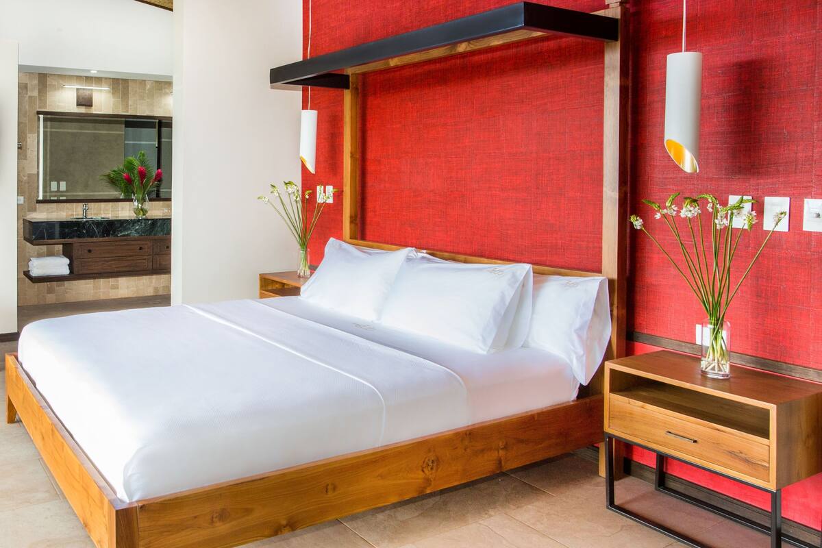 A modern hotel room with a wooden bed, a red accent wall, a wooden nightstand, and a bathroom with a marble countertop.