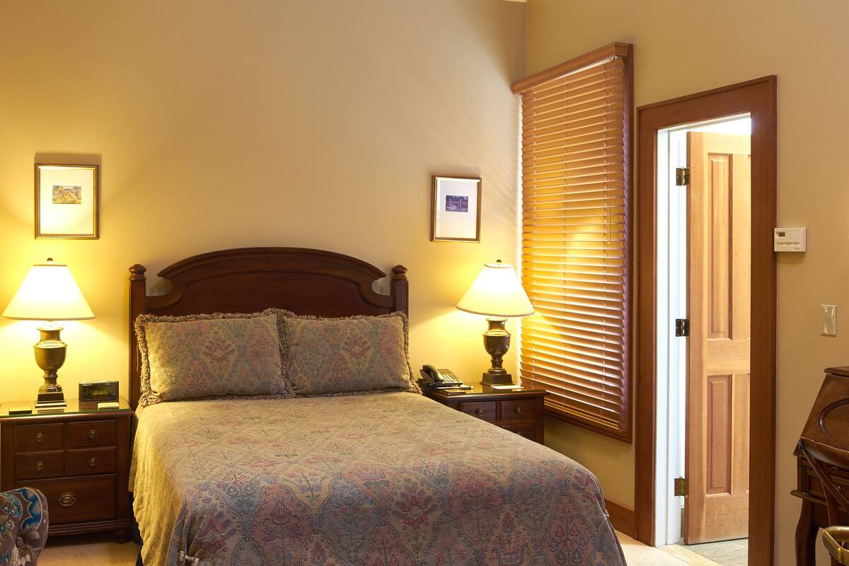 A bedroom with a large bed, two bedside tables with lamps, a chair, a small ottoman, a window with blinds, and a door.