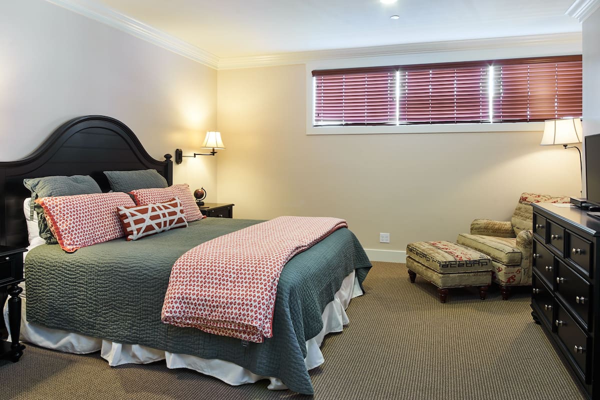 A neatly arranged bedroom with a bed, bedside lamps, a window with blinds, a chair, and a dresser.